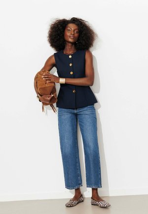 Femme aux cheveux bouclés portant un haut sans manches bleu marine, un jean bleu, des ballerines imprimé léopard, tenant un sac à dos en daim marron contre un mur blanc.