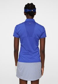 Blå kortärmad polo med en mesh baksidedesign och logotyp på kragen, i kombination med en mönstrad vit och blå kjol.