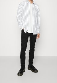 Camisa de botones de rayas blancas y azul marino sobre una camiseta blanca sencilla, combinada con jeans ajustados negros y zapatos negros de cordones con costuras amarillas.
