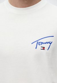 Λευκό μπλουζάκι με μπλε λογότυπο "Tommy" σε χειρόγραφη γραφή και μικρό ορθογώνιο κόκκινο, άσπρο και μαύρο έμβλημα με σημαία στο στήθος.