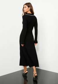 Schwarzes geripptes Strickkleid mit langen Ärmeln und schwingendem Saum. Verfügt über einen kontrastierenden weißen Kragen und eine taillierte Taille. Das Modell trägt schwarze Keilabsätze.