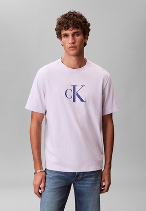Hombre con cabello rizado y castaño que lleva una camiseta de color rosa pálido con el logo de Calvin Klein y jeans azules, sobre un fondo gris liso.