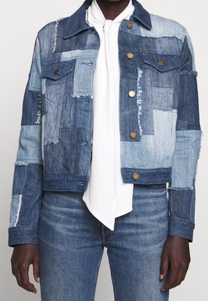 Personne portant une veste en denim patchwork avec des boutons en laiton, tons bleu clair et foncé, associée à un jean délavé moyen et une écharpe blanche nouée autour du cou.