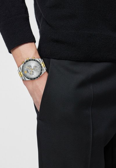 Randmekell hõbedase ja kulla metallrihmaga, hõbedane kellanägu koos kronograafiga, kantud vasakul randmel musta kampsuni ja mustade pükstega inimese käel.