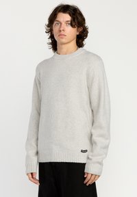Lys grå, genser med rund hals laget av mykt materiale med ribbestrikkede mansjetter og hem; har et lite svart logomerke på undersiden.