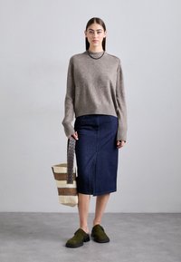 Nainen, jolla on päällään beige neule, tummansiniset polvipituinen hame, oliivinvihreät kengät, ja hän pitää käsissään raitakuosisia kudottua laukkua, jonka kahva on nahkaa.