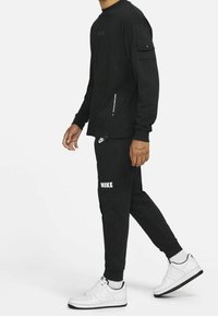 Musta Nike pitkähihainen paita, jossa on pyöreä kaula-aukko ja sivutasku. Mustat Nike-collegehousut logolla. Valkoiset Nike-tennarit mustalla pohjalla.