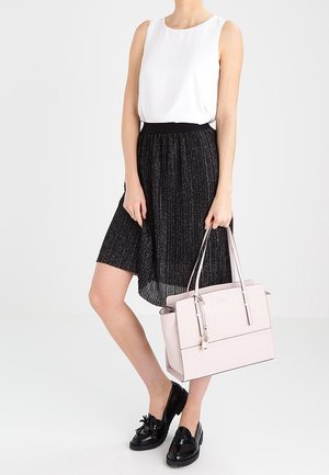 Donna che indossa un top bianco senza maniche, una gonna nera a pieghe, mocassini neri, mentre tiene una borsa strutturata rosa chiaro per i manici.