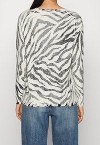 Pull en tricot blanc avec des rayures zèbre noires, présentant un ourlet festonné et un col rond, associé à un jean en denim bleu.