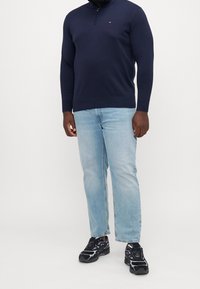 Homme portant un pull quart-zip bleu marine, un jean bleu clair et des chaussures de sport noires, debout devant un fond uni.