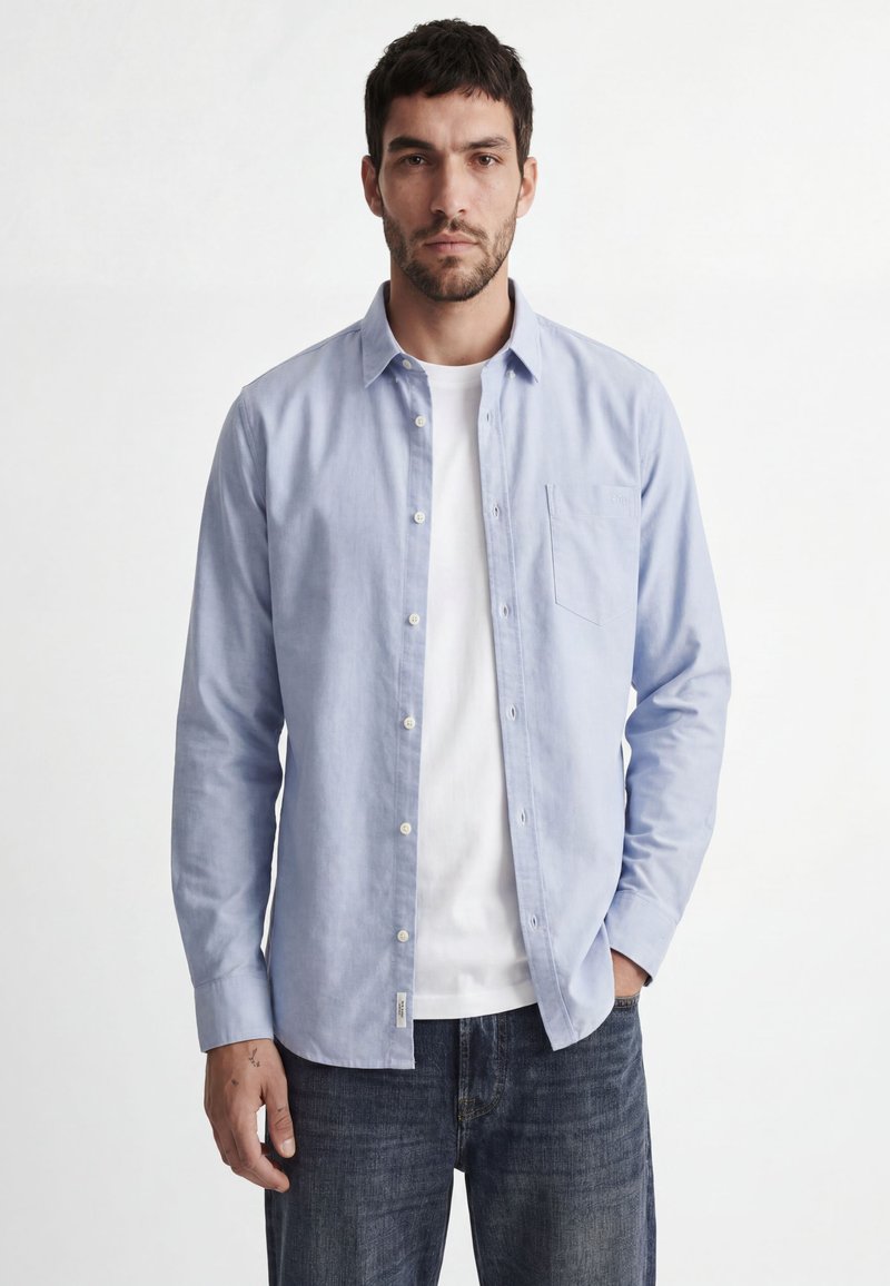 Mann mit dunklem Haar und Bart, trägt ein hellblaues Hemd über einem weißen T-Shirt und dunklen Jeans vor einfarbig weißem Hintergrund.