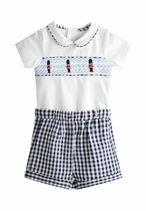 Biały kombinezon dla niemowląt z haftowanymi królewskimi strażnikami, kołnierzykiem typu Peter Pan oraz dołączonymi granatowymi szortami w kratę gingham.