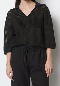 Schwarzer, offener Strickpullover mit V-Ausschnitt und einem kragenartigen, strukturierten Halsausschnitt. Mit 3/4-Ärmeln und einem lässigen Schnitt versehen. Kombiniert mit schwarzen Hosen.
