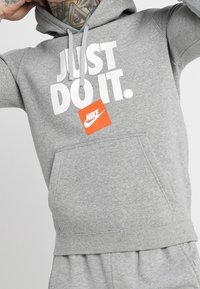 Sudadera gris con "JUST DO IT." impreso en blanco y una etiqueta del logo de Nike en naranja. Cuenta con un bolsillo frontal y capucha con cordón. Tejido de textura suave.