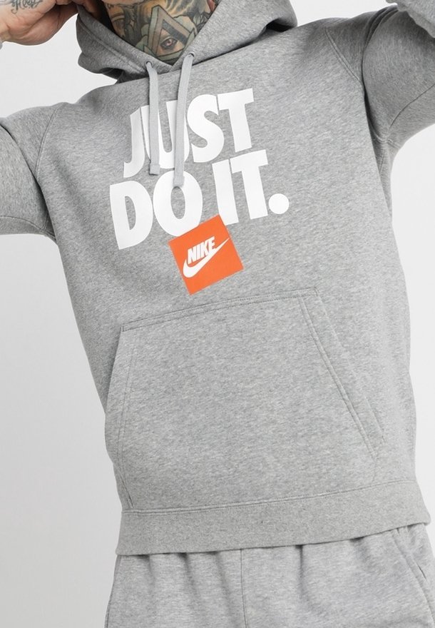 Sudadera gris con "JUST DO IT." impreso en blanco y una etiqueta del logo de Nike en naranja. Cuenta con un bolsillo frontal y capucha con cordón. Tejido de textura suave.