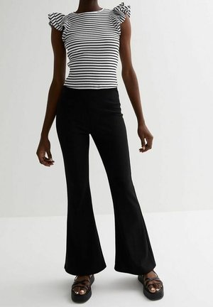 Kvinde iført sorte vidde bukser, sorte platformsandaler og en hvid kortærmet top med sorte vandrette striber og flæseærmer.