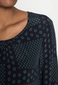 Blusa blu navy con un motivo testurizzato di cerchi e quadrati verdi, dotata di un collo arrotondato e di delicati arricciamenti sulla spalla.