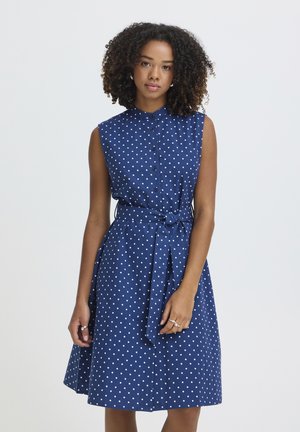 Vrouw met krullend haar, gekleed in een mouwloze marineblauwe jurk met witte polka dots, een knoopsluiting aan de voorkant en een ceintuur in de taille.