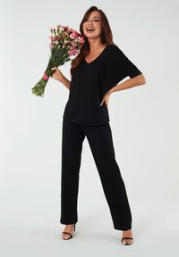 Czarna bluzka z krótkim rękawem i dekoltem w serek, zestawiona z czarnymi spodniami, połączona z czarnymi sandałami na wysokim obcasie. Modelka trzyma bukiet różowych kwiatów.