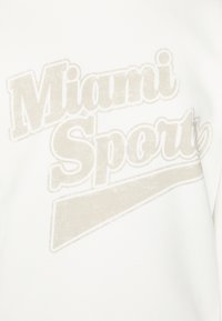 Vit sweatshirt med en bleknad kr ämfärgad grafik som har texten "Miami Sport" i ett retro typsnitt, accentuerad av ett diagonalt designelement.