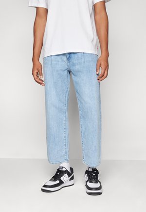 Jean en denim bleu clair, ample et à coupe droite, avec des ourlets retroussés. Associé à des baskets noir et blanc et un t-shirt blanc.