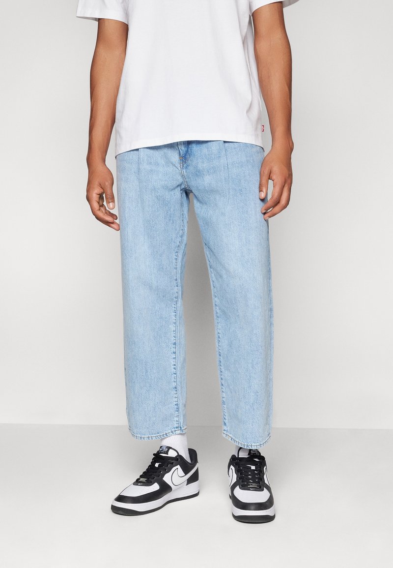 Jean en denim bleu clair, ample et à coupe droite, avec des ourlets retroussés. Associé à des baskets noir et blanc et un t-shirt blanc.