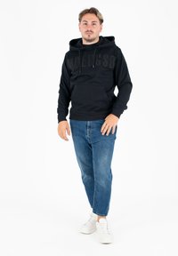 Felpa nera con tasca a marsupio frontale e design testuale in rilievo, abbinata a jeans blu e sneakers bianche, in piedi su uno sfondo bianco.