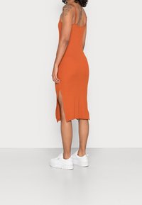 Robe ajustée orange avec fines bretelles, fentes latérales et ourlet à hauteur des genoux. Assortie à des baskets blanches. Texture lisse.