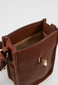 Piccolo borsetta a tracolla in pelle marrone con chiusura in tono oro, foderata in beige e con tracolla regolabile, su sfondo bianco semplice.