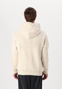 Sudadera beige con un gran logotipo tonal en la espalda, que cuenta con una capucha ajustable y puños y dobladillo acanalados. Textura de tela suave.