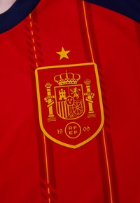 Κόκκινη ποδοσφαιρική φανέλα με κίτρινο έμβλημα στέμματος που έχει τη λέξη "España", το έτος "1909" και επιπλέον σχέδια που περιλαμβάνουν κατακόρυφες ρίγες.