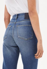 Modré džíny z denimu s vysokým pasem, designem s pěti kapsami a koženou nášivkou s logem vzadu. Vyznačují se kontrastním šitím a hladkou texturou.