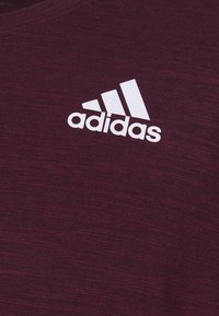Valkoinen Adidas-logo, jossa on kolme diagonaalista raitaa, tumman viininpunaisella teksturoidulla kangastaustalla.