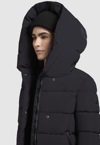 Eine Frau trägt eine schwarze Strickmütze und eine schwarze Daunenjacke mit einer großen Kapuze vor einem schlichten, hellen Hintergrund.