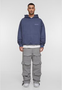 Marineblå hettegenser med kengurulomme og preget logo, sammen med lysegrå cargo-bukser med flere lommer og en avslappet passform. Hvite sneakers.