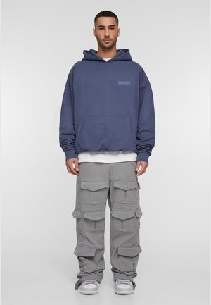 Marineblå hettegenser med kengurulomme og preget logo, sammen med lysegrå cargo-bukser med flere lommer og en avslappet passform. Hvite sneakers.