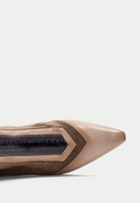 Zapato de tacón de punta con una mezcla de cuero marrón claro y ante marrón; presenta una sección central texturizada con un detalle de cremallera transparente.