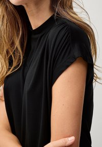 Sort kortærmet top med høj halsudskæring. Stoffet ser blødt ud og falder løst med subtil rynkning ved skuldrene.