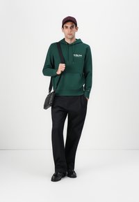 Sweatshirt verde com capuz e bolso frontal, calças largas pretas e sapatos pretos. Os acessórios incluem uma bolsa cinza e um boné bordô.