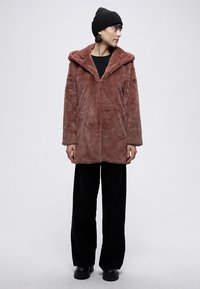 Mākslīgās kažokādas mētelis putekļainā rožu krāsā; tam ir plats apkaklis, gari piedurkņi un divas priekšējās kabatas. Valkāts kopā ar melnām biksēm un apaviem.