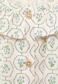 Tessuto crema con motivo esagonale che presenta piccoli fiori blu e foglie verdi, decorato con bottoni in legno marrone.