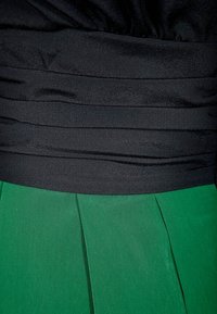 Corsage noir ajusté avec des plis horizontaux se transformant en une jupe verte fluide à plis verticaux, mettant en valeur des textures et des couleurs contrastées.