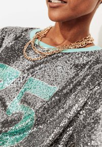 Pull argenté à sequins avec un col vert menthe et un grand motif "35". Trois rangs de colliers en chaîne dorée sont visibles.