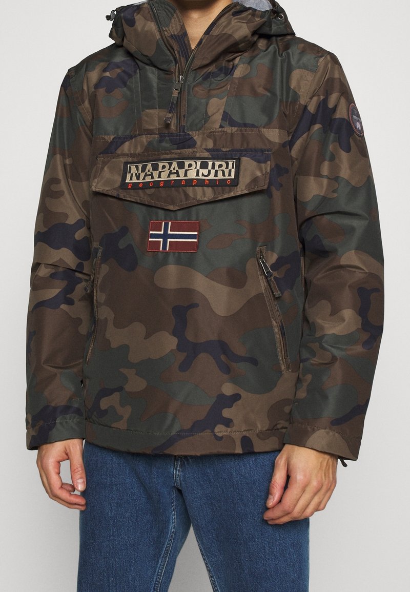 Homme portant une veste à capuche camouflage marron et verte avec une poche ventrale, des détails zippés et un patch de drapeau sur la poitrine.