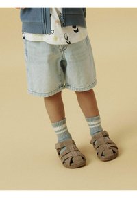 Sandalias de ante marrón con tiras abiertas, combinadas con calcetines a rayas azules. El niño lleva pantalones cortos de mezclilla claros y una camisa estampada.