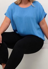 Femme assise contre un mur blanc, portant un chemisier bleu à manches courtes ample et un pantalon noir ajusté, avec des cheveux blonds visibles.