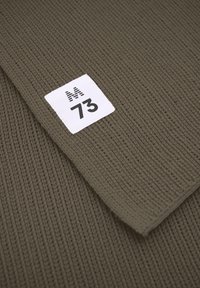 Nærbillede af brun strikket stof med et hvidt firkantet label, der viser "M 73" syet i hjørnet.