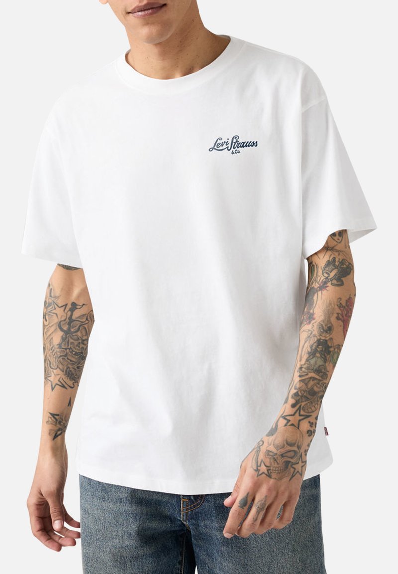 Hombre con camiseta blanca de Levi's con pequeño logo en el pecho y brazos fuertemente tatuados, combinado con vaqueros azules.