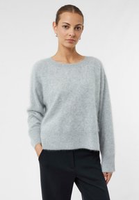 Jasnoszary sweter wykonany z miękkiego, puszystego materiału; ma okrągły dekolt, długie rękawy i prążkowany dół. W zestawie z ciemnymi spodniami.
