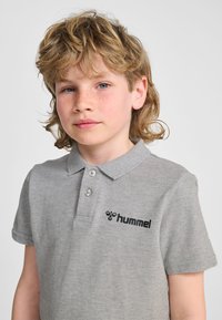 Szara koszulka polo wykonana z miękkiego materiału, z kołnierzykiem, trzema guzikami oraz czarnym logo „hummel” po lewej stronie.